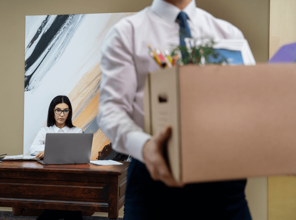 Employee leaving office with a box of belongings while HR manager works on paperwork