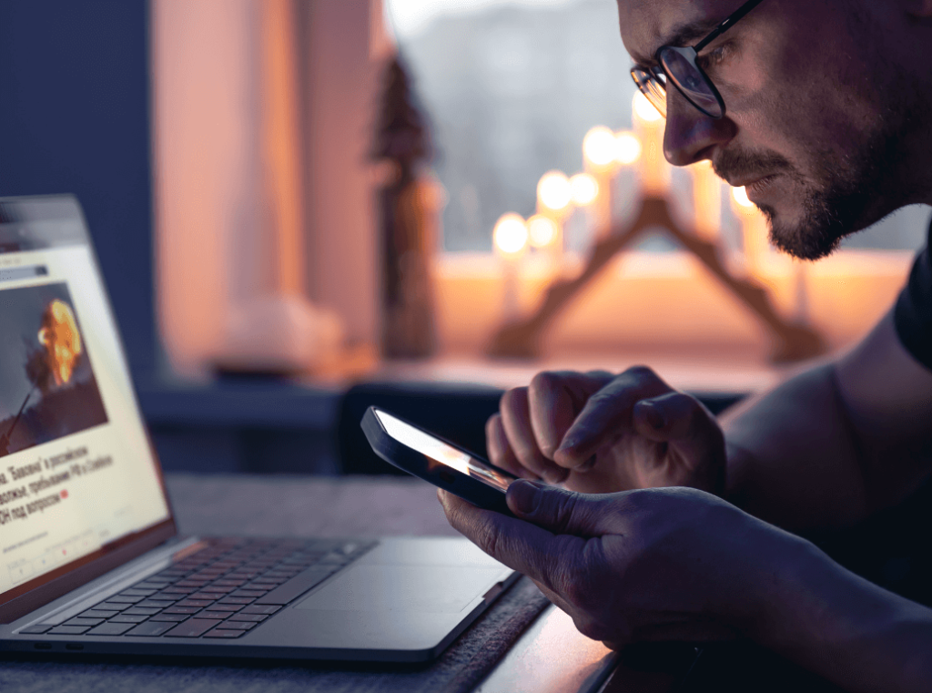 A close-up of a professional using a smartphone and laptop simultaneously to review online content and digital footprints.