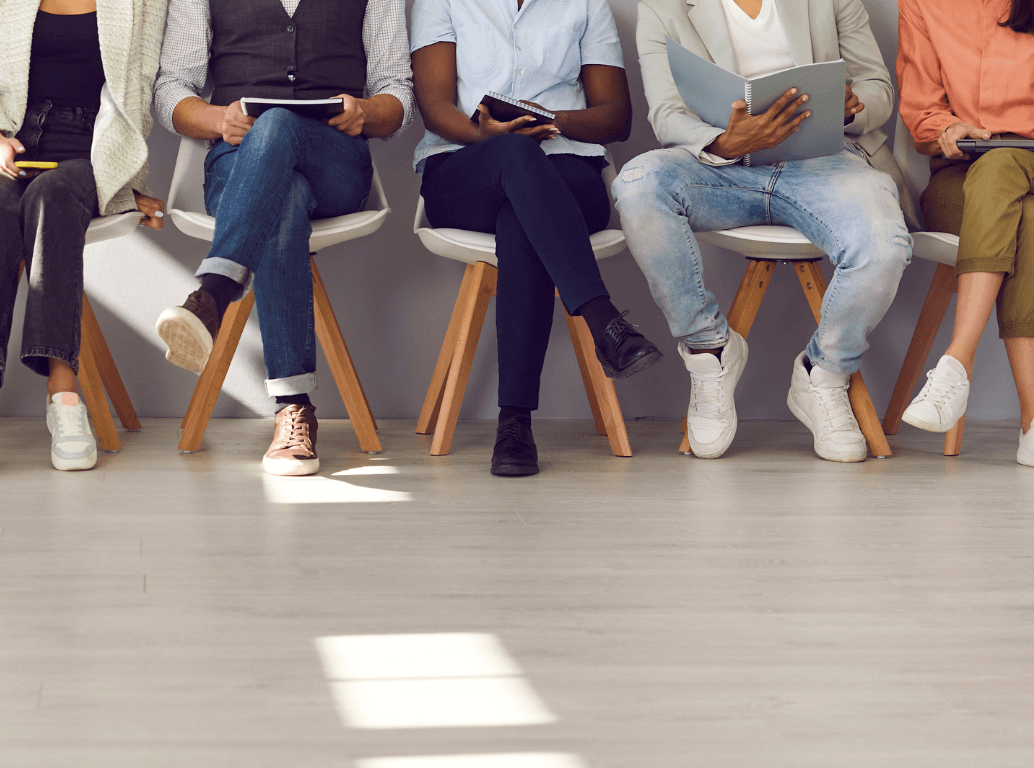 A row of diverse candidates sitting side-by-side in chairs holding notebooks, waiting for their job interviews during a peak recruitment season.