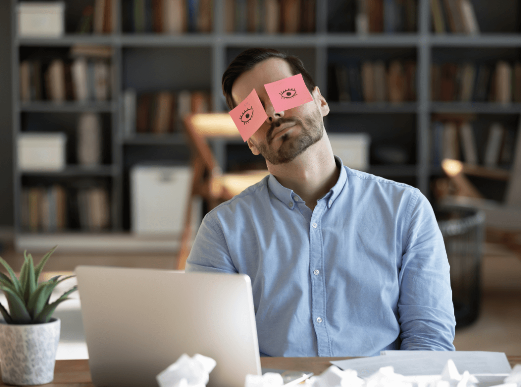 An employee slacking off at his desk, wearing sticky notes with drawn eyes to pretend he is working, illustrating the risk of hiring poor performers.