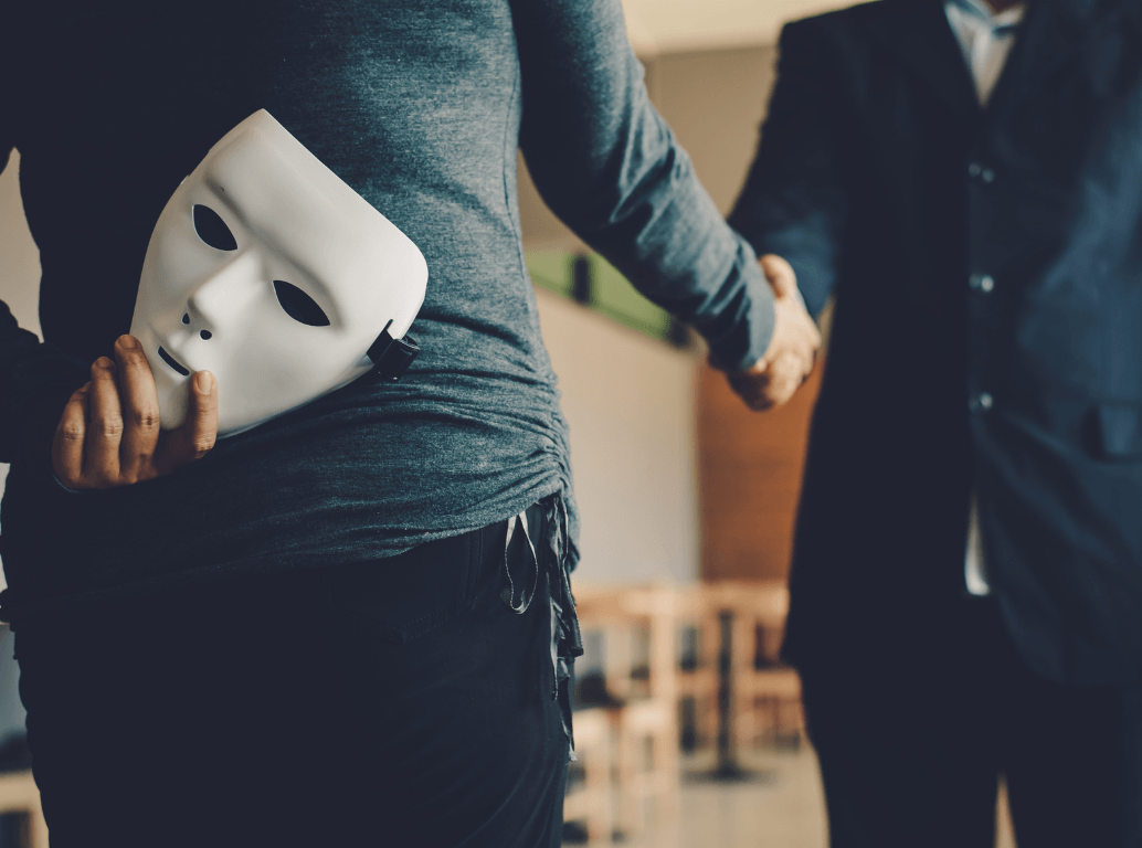 A person holding a white mask behind their back while shaking hands with an executive, representing hidden deception and the need for executive background checks.