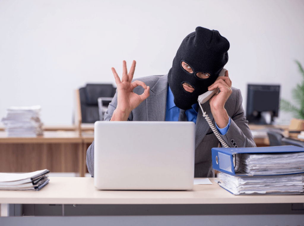 A man in a business suit wearing a black balaclava while working on a laptop, symbolising the internal risk of corporate fraud and white-collar crime.
